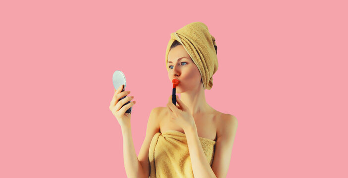 Portrait of beautiful young woman painting her lips with red lipstick applying makeup looking in pocket mirror and drying her wet hair with wrapped bath towel on her head on pink background