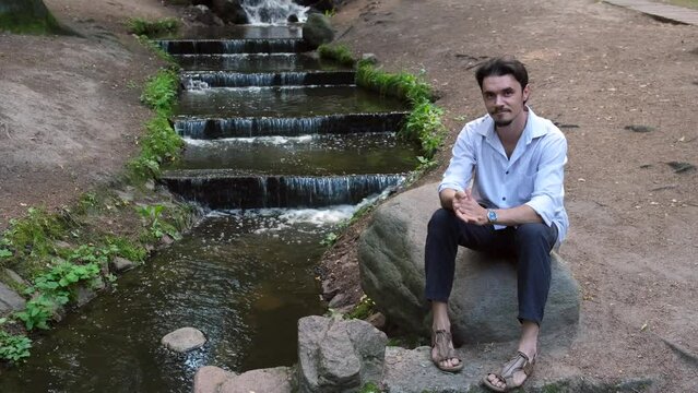 Young handsome man sits on stone near water in park. Looks at camera and smiles. Man enjoys calmness, murmur of water unity with nature. Outdoors. Happiness.4k resolution footage, film, moving image. 
