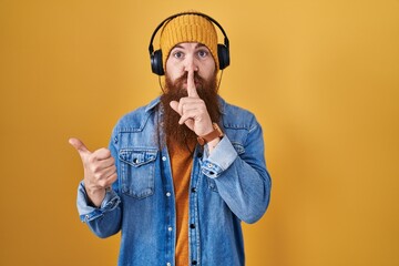 Caucasian man with long beard listening to music using headphones asking to be quiet with finger on lips pointing with hand to the side. silence and secret concept.