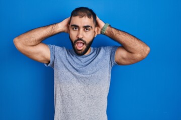 Middle east man with beard standing over blue background crazy and scared with hands on head,...