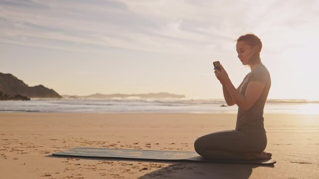Woman scrolling her smartphone on ocean beach at sunset. Female sitting on Yoga mat on sand sea shore at sunrise. Woman checking exercises online with phone in the morning, evening, outdoors