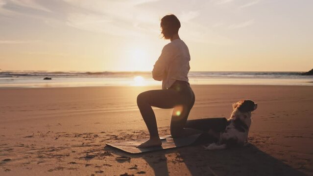 Woman doing workout with dog on ocean beach at sunset. Female doing Yoga and stretching on mat on sand sea shore at sunrise. Strong powerful woman exercises in the morning or evening outdoors. 