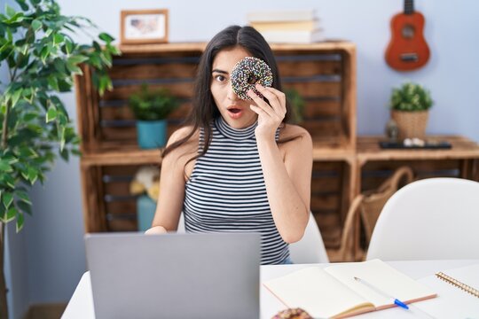 Young teenager girl using computer laptop with doughnut on face scared and amazed with open mouth for surprise, disbelief face