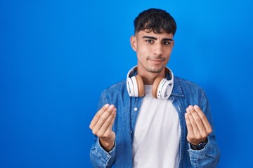 Young hispanic man standing over blue background doing money gesture with hands, asking for salary...