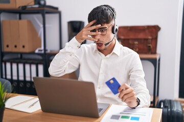 Young hispanic man working using computer laptop holding credit card peeking in shock covering face and eyes with hand, looking through fingers with embarrassed expression.