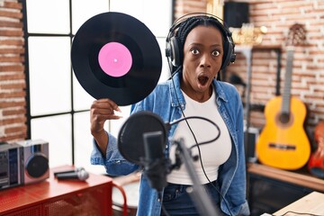 Beautiful black woman holding vinyl record at music studio scared and amazed with open mouth for surprise, disbelief face