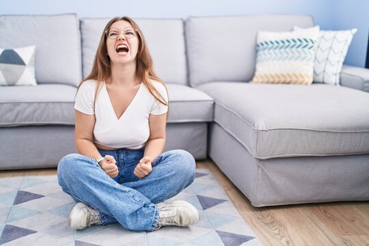 Young Caucasian Woman Sitting On The Floor At The Living Room Angry And Mad Screaming Frustrated And Furious, Shouting With Anger. Rage And Aggressive Concept.