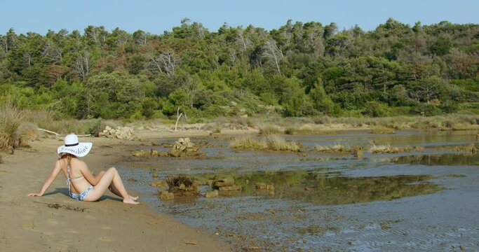 A young woman with a white hat is sitting on a sand beach, Rab Island, Croatia   