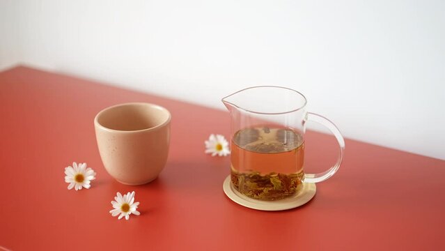 Close-up of a glass teapot filled with herbal tea and a ceramic minimalist mug and chamomile flower buds on red furniture. Stress relief