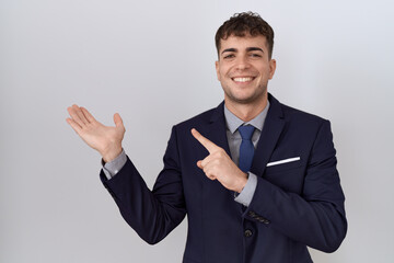 Young hispanic business man wearing suit and tie amazed and smiling to the camera while presenting with hand and pointing with finger.