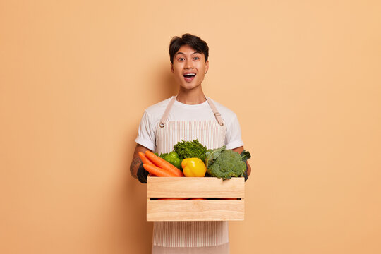 Young farmer stands with box of vegetables on peachy background in studio, smiles joyfully, organic food concept, copy space