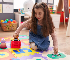 Adorable hispanic girl playing with tractor toy sitting on floor at kindergarten