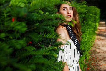 Side view of beautiful female with red lipstick in white off-the-shoulder dress pressing back to pine tree and looking at camera with slightly open mouth.