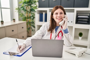 Young blonde woman doctor talking on telephone writing on document at clinic