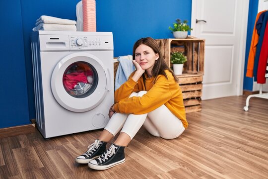 Young blonde woman sitting on floor waiting for washing machine smiling at laundry room