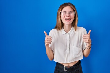 Beautiful woman standing over blue background success sign doing positive gesture with hand, thumbs up smiling and happy. cheerful expression and winner gesture.