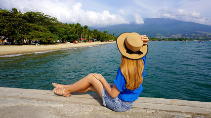 Beautiful young woman with hat sitting on pier looking at panoramic view of Ilhabela Island, Brazil