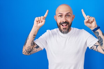 Naklejka premium Hispanic man with tattoos standing over blue background smiling amazed and surprised and pointing up with fingers and raised arms.