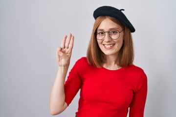 Young redhead woman standing wearing glasses and beret showing and pointing up with fingers number three while smiling confident and happy.