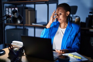 Beautiful african american woman working at the office at night smelling something stinky and disgusting, intolerable smell, holding breath with fingers on nose. bad smell