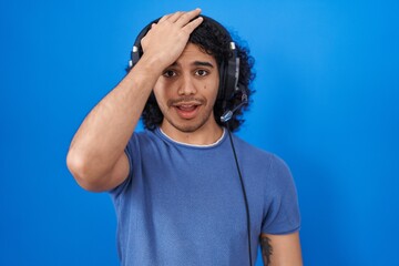 Hispanic man with curly hair listening to music using headphones surprised with hand on head for mistake, remember error. forgot, bad memory concept.