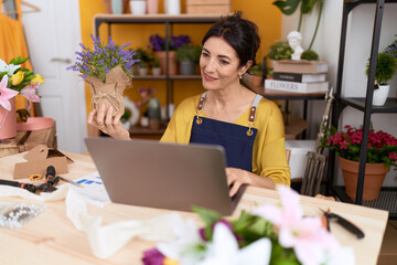 Middle age hispanic woman florist using laptop holding lavender plant at flower shop