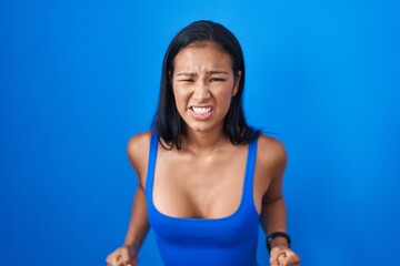 Fototapeta na wymiar Hispanic woman standing over blue background angry and mad screaming frustrated and furious, shouting with anger. rage and aggressive concept.