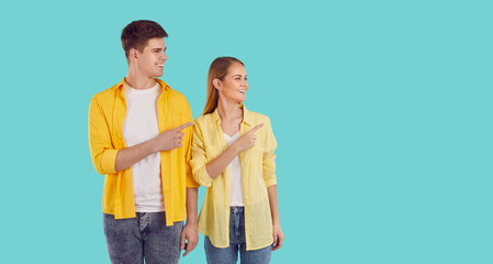Young attractive Caucasian man and woman pointing finger to side giving you recommendation smiling looking at copy space for advertising dressed in jeans and casual shirt stands in turquoise studio