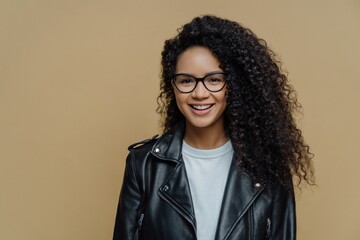 Stylish hipster girl, bushy Afro hair, positive smile, enjoying day, glasses, black leather jacket. Brown background. People, emotions, fashion concept.