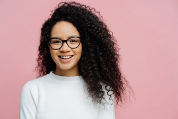 Charming young female, Afro hairstyle, pleasant smile, very happy. Transparent glasses, white jumper. Isolated on rosy background. Amused ethnic female.