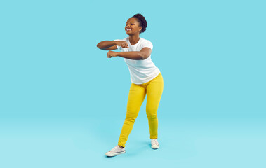 Fototapeta na wymiar Young attractive African American woman performs funny dance enjoying life or make simple daily gymnastics dressed in white casual t-shirt and trousers, standing in turquoise studio