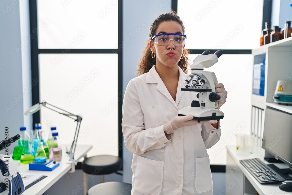 Sticker young hispanic woman working at scientist laboratory puffing cheeks with funny face. mouth inflated 