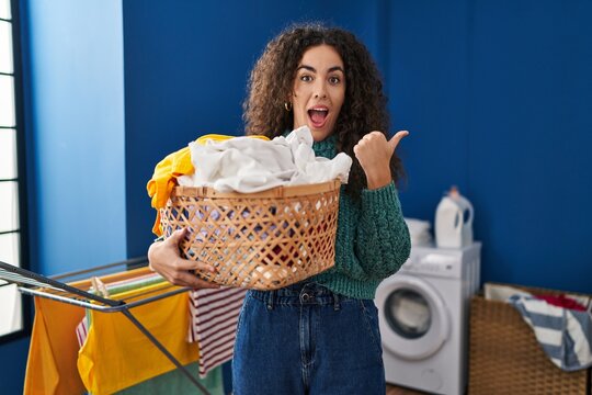 Young hispanic woman holding laundry basket pointing thumb up to the side smiling happy with open mouth