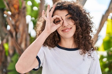 Hispanic woman with curly hair standing outdoors smiling happy doing ok sign with hand on eye looking through fingers