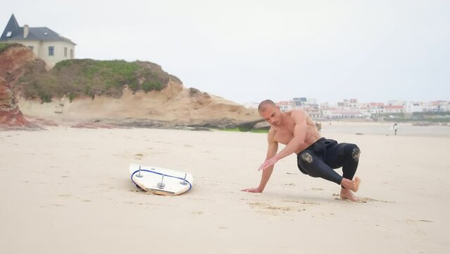 Young man with athletic body do warm-up before surfing on the beach near the ocean. Surfer in wetsuit doing active exercises to prepare the body for surf. Guy do stretching movements in sand outdoors