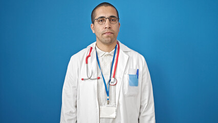 Fototapeta premium Young hispanic man doctor standing with serious expression over isolated blue background