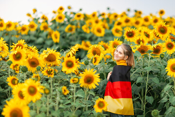 Teen girl holding flag of Germany on her shoulders in the sunflower field. Day of German Unity. Travel and learn german language concept. Selective focus.