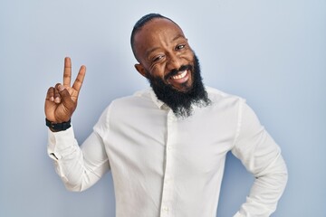 African american man standing over blue background smiling looking to the camera showing fingers doing victory sign. number two.