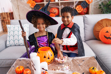Fototapeta premium Adorable african american boy and girl wearing halloween costume putting candies in pumpkin basket at home
