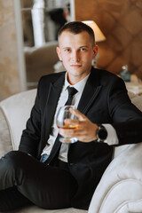 a young man sitting in a chair with a glass of alcoholic drink in a hotel room, a young businessman wearing a black suit and a white shirt with a tie. The groom is preparing for the wedding ceremony.