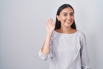 Young hispanic woman standing over white background waiving saying hello happy and smiling,...