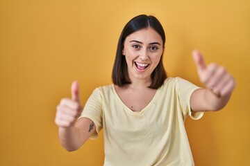 Hispanic girl wearing casual t shirt over yellow background approving doing positive gesture with hand, thumbs up smiling and happy for success. winner gesture.