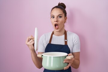 Young hispanic girl wearing apron holding cooking pot afraid and shocked with surprise and amazed...