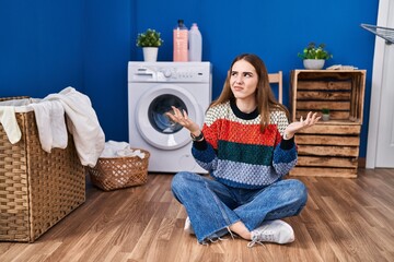 Young hispanic girl doing laundry clueless and confused expression with arms and hands raised. doubt concept.