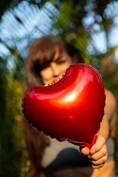 close em balão de coração vermelho metalizado sendo oferecido por uma mulher morena com ela e o fundo de natureza desfocados.