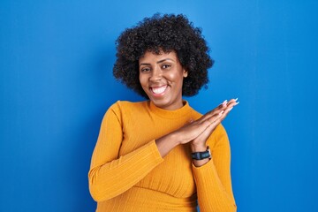 Black woman with curly hair standing over blue background clapping and applauding happy and joyful,...