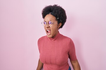 Fototapeta na wymiar Beautiful african woman with curly hair standing over pink background angry and mad screaming frustrated and furious, shouting with anger. rage and aggressive concept.