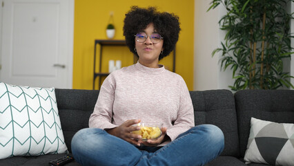 Young african american woman watching tv sitting on sofa eating chips at home