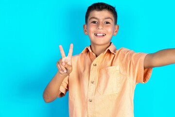 Positive Little hispanic boy wearing orange shirt take selfie make v-sign