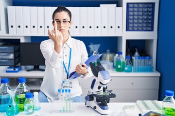 Young brunette woman working at scientist laboratory showing middle finger, impolite and rude fuck...
